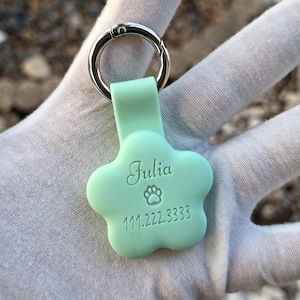 Könnte beinhalten: Ein hellgrüner Haustieranhänger in Form eines Pfotenabdrucks. Der Anhänger trägt den Namen "Julia", ein Pfotenabdrucksymbol und eine Telefonnummer. Er ist an einem silbernen Metallring befestigt.