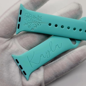 Può includere: Due cinturini per Apple Watch di colore turchese, uno con un design floreale e l'altro con il nome "Kayla" inciso.
