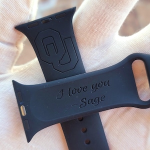 May include: Black silicone watch band with a custom engraved message "I love you - Sage" and a University of Oklahoma logo.