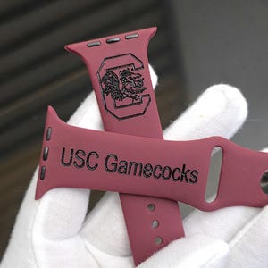 May include: Burgundy-colored watch band with the text "USC Gamecocks" and a Gamecock logo. The band is held by a gloved hand. The band is made of a flexible material.