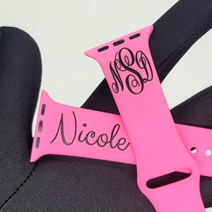 May include: Two pink Apple Watch bands with black engraved text. One band says "Nicole" and the other has the monogram "NSD".