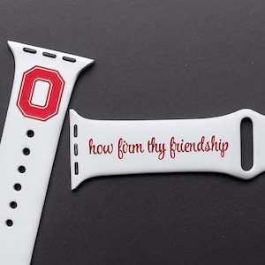 May include: Two white Apple Watch bands. One band features a red and white "O" logo. The other band has the phrase "how firm thy friendship" in red script. The bands are on a dark gray background.