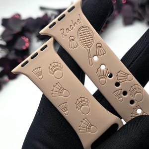 Puede incluir: Una banda beige para Apple Watch con temática de bádminton. La banda tiene un diseño de raqueta de bádminton y volante blancos. La banda está personalizada con el nombre "Rachel".