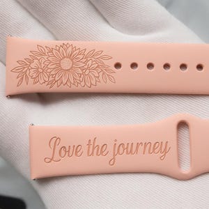 Può includere: Due cinturini per orologio rosa chiaro. Uno presenta un design floreale con un girasole e altri fiori. L'altro cinturino ha incisa la frase "Love the journey". I cinturini sono realizzati in un materiale morbido e flessibile.