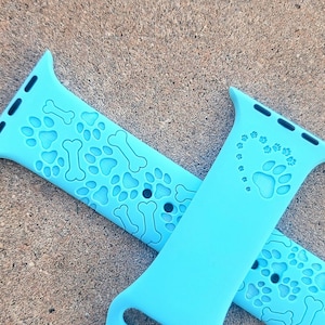 Puede incluir: Dos correas para Apple Watch de color azul claro con diseños de huellas de patas y huesos. Una correa presenta un corazón en forma de huellas de patas.