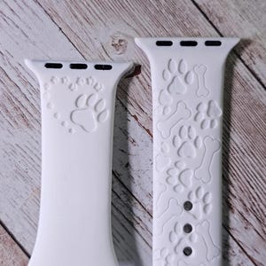 Puede incluir: Dos correas de reloj de silicona blancas con diseños de huellas de patas y huesos. La correa de la izquierda tiene una sola huella de pata en el centro, mientras que la correa de la derecha tiene un patrón repetido de huellas de patas y huesos.