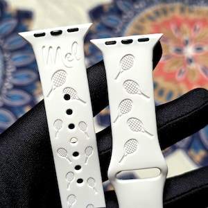 Puede incluir: Dos correas de reloj blancas con diseños de raquetas de tenis. Una correa tiene el nombre "Mel" grabado. Las correas tienen agujeros de ajuste y se muestran sobre un fondo estampado.