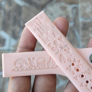 May include: Two pink silicone watch bands with white embossed Disney characters and designs. One band features a castle, Mickey Mouse, and other icons. The other band features a tree, a roller coaster, and a castle.