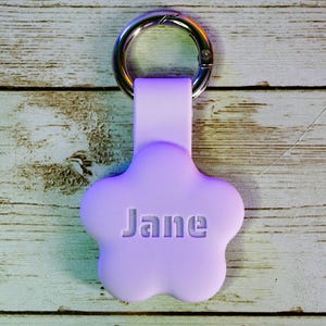 May include: A purple flower-shaped keychain with a silver ring and the name "Jane" embossed on the front.