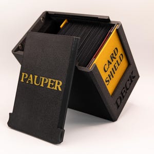 Könnte beinhalten: Schwarze Kartenaufbewahrungsbox mit einem gelben Etikett mit der Aufschrift "CARD SHIELD DECK" und einem schwarzen Deckel mit einem gelben Etikett mit der Aufschrift "PAUPER".