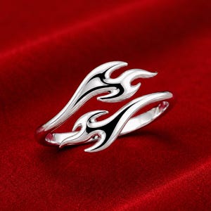May include: A silver-toned open ring featuring a stylized flame design. The band splits into two tapered ends, each shaped like a flickering flame with smooth, polished surfaces and dark recessed detailing to define the flame contours.