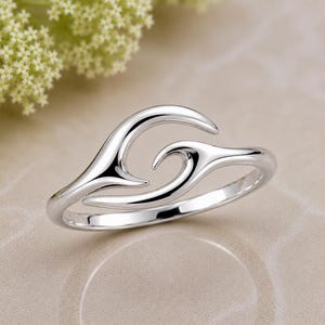 May include: A polished silver ring featuring a continuous, fluid design that mimics the shape of a cresting wave. The band is smooth and reflective, with two tapered ends that curve inward toward each other to create an open, abstract wave motif.