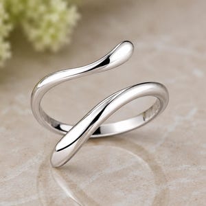 Peut inclure: Une bague ouverte en métal argenté poli présentant un design fluide, lisse et continu en forme de vague. L'anneau se courbe en deux extrémités effilées qui se chevauchent légèrement, créant une surface métallique lisse et réfléchissante.