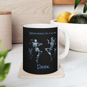 May include: White ceramic mug with a black background and a blue graphic of skeletons dancing. The text "Radiologists do it in the dark" is printed in blue on the mug.