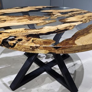 K&ouml;nnte beinhalten: Runder Holztisch mit einer durchsichtigen Epoxidharzplatte und einem schwarzen Metallgestell. Die Tischplatte hat eine nat&uuml;rliche Holzmaserung und das Harz hat ein flussartiges Design.
