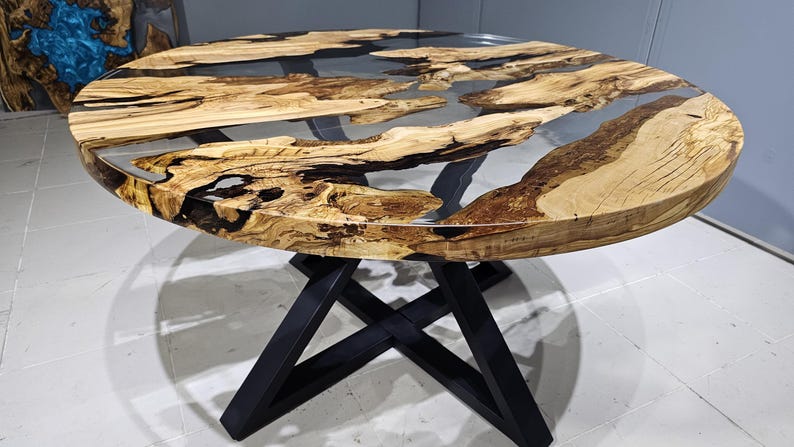 K&ouml;nnte beinhalten: Runder Holztisch mit einer durchsichtigen Epoxidharzplatte und einem schwarzen Metallgestell. Die Tischplatte hat eine nat&uuml;rliche Holzmaserung und das Harz hat ein flussartiges Design.