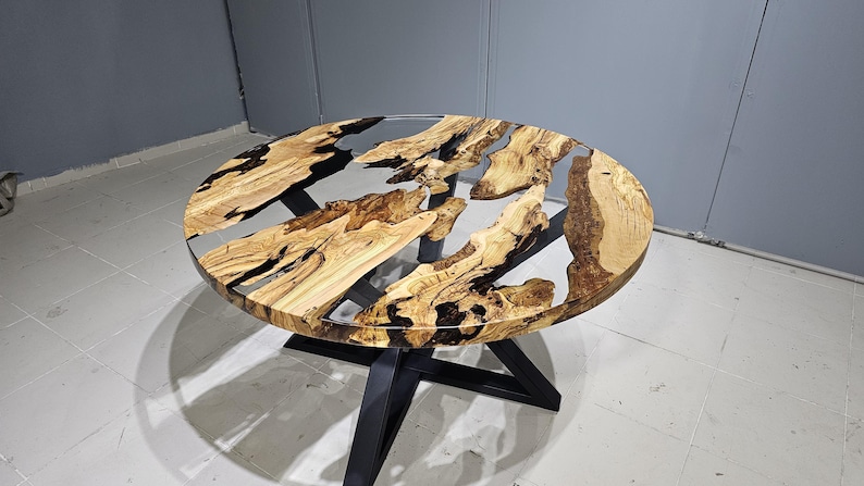 K&ouml;nnte beinhalten: Runder Holztisch mit einem schwarzen Metallgestell. Die Tischplatte besteht aus Holz, &uuml;ber das klares Epoxidharz gegossen wurde, wodurch ein einzigartiges Flussdesign entsteht.