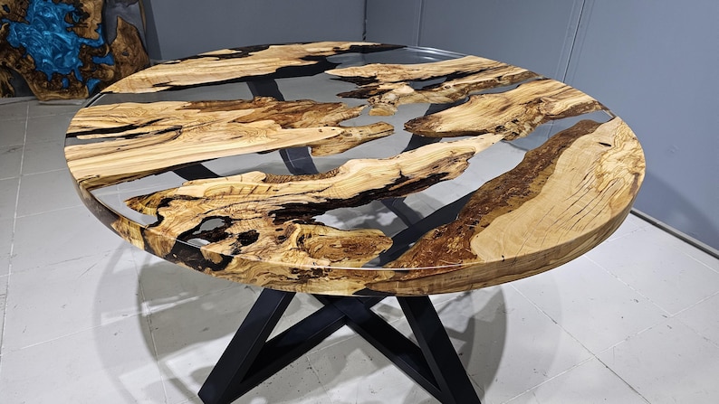 K&ouml;nnte beinhalten: Runder Holztisch mit einer durchsichtigen Epoxidharzplatte. Der Tisch hat ein nat&uuml;rliches Holzmaserungsmuster und schwarze Epoxidharzakzente. Der Tisch wird von einem schwarzen Metallgestell getragen.