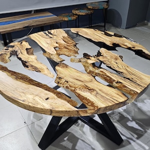K&ouml;nnte beinhalten: Runder Holztisch mit schwarzem Metallgestell. Die Tischplatte besteht aus Naturholz mit einem klaren Epoxidharzguss, der ein flussartiges Design mit schwarzen und wei&szlig;en Wirbeln erzeugt.