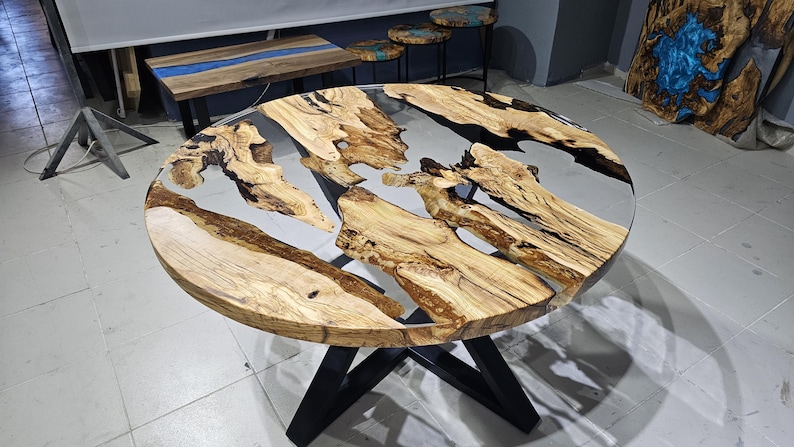 K&ouml;nnte beinhalten: Runder Holztisch mit schwarzem Metallgestell. Die Tischplatte besteht aus Naturholz mit einem klaren Epoxidharzguss, der ein flussartiges Design mit schwarzen und wei&szlig;en Wirbeln erzeugt.