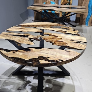 K&ouml;nnte beinhalten: Runder Holztisch mit schwarzem Metallgestell. Die Tischplatte besteht aus Holz mit einer klaren Epoxidharz-Oberfl&auml;che. Das Harz hat ein nat&uuml;rliches, flie&szlig;endes Muster.