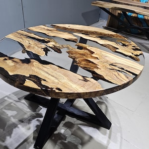 K&ouml;nnte beinhalten: Runder Holztisch mit einer schwarzen Epoxidharzplatte und einem schwarzen Metallgestell. Die Tischplatte hat eine nat&uuml;rliche Holzmaserung und das Harz hat eine klare, glasartige Oberfl&auml;che.
