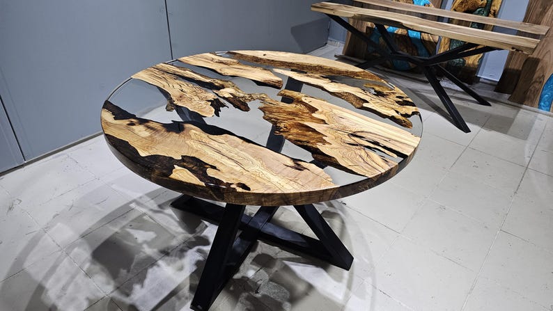 K&ouml;nnte beinhalten: Runder Holztisch mit einer schwarzen Epoxidharzplatte und einem schwarzen Metallgestell. Die Tischplatte hat eine nat&uuml;rliche Holzmaserung und das Harz hat eine klare, glasartige Oberfl&auml;che.