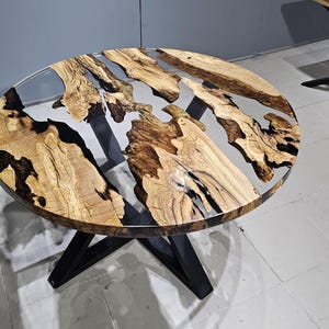 K&ouml;nnte beinhalten: Runder Holztisch mit einer durchsichtigen Epoxidharzplatte. Der Tisch hat ein nat&uuml;rliches Holzmaserungsmuster und schwarze Epoxidharz-Akzente. Der Tisch wird von einem schwarzen Metallgestell getragen.