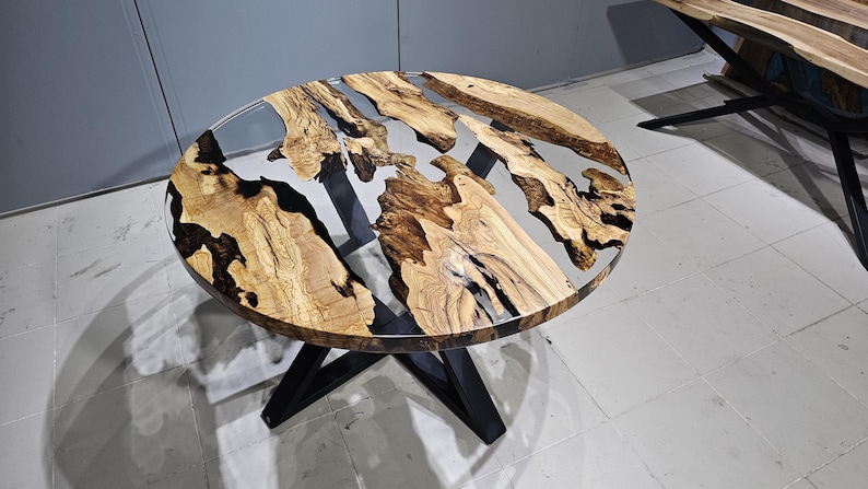 K&ouml;nnte beinhalten: Runder Holztisch mit einer durchsichtigen Epoxidharzplatte. Der Tisch hat ein nat&uuml;rliches Holzmaserungsmuster und schwarze Epoxidharz-Akzente. Der Tisch wird von einem schwarzen Metallgestell getragen.
