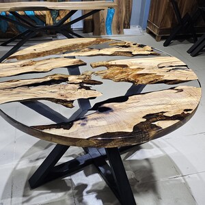 K&ouml;nnte beinhalten: Runder Holztisch mit einer durchsichtigen Epoxidharzplatte. Der Tisch hat ein schwarzes Metallgestell mit einem einzigartigen X-f&ouml;rmigen Design.