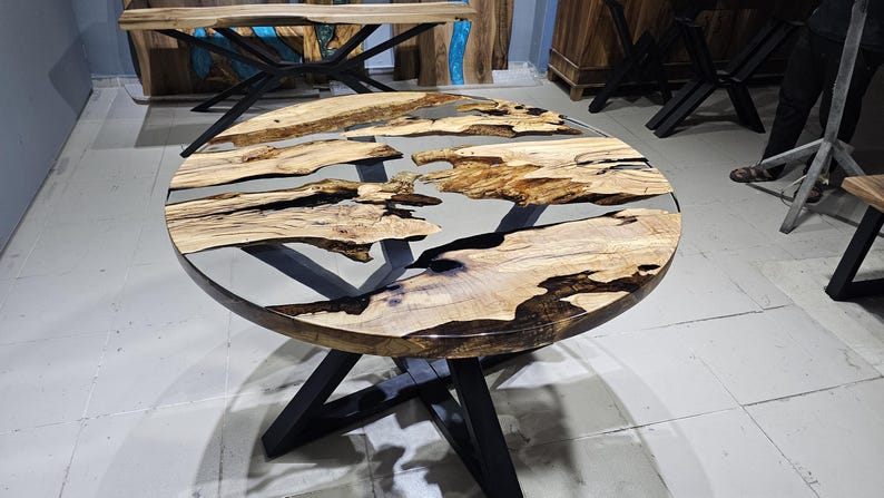 K&ouml;nnte beinhalten: Runder Holztisch mit einer durchsichtigen Epoxidharzplatte. Der Tisch hat ein schwarzes Metallgestell mit einem einzigartigen X-f&ouml;rmigen Design.