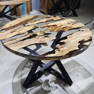 K&ouml;nnte beinhalten: Runder Holztisch mit einem schwarzen Metallgestell. Die Tischplatte besteht aus Holz mit einem klaren Epoxidharzguss, der ein flussartiges Design mit schwarzen und blauen Akzenten erzeugt.