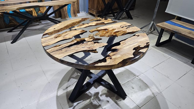 K&ouml;nnte beinhalten: Runder Holztisch mit einem schwarzen Metallgestell. Die Tischplatte besteht aus Holz mit einem klaren Epoxidharzguss, der ein flussartiges Design mit schwarzen und blauen Akzenten erzeugt.