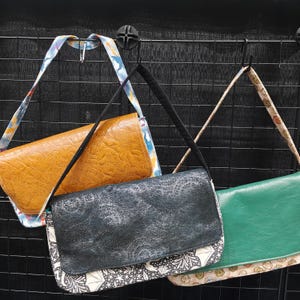 May include: Three rectangular flap bags hanging on a black wire grid. The left bag is textured mustard yellow with a blue and white patterned strap. The center bag is black with a textured flap and a black strap. The right bag is green with a patterned strap.