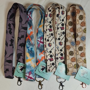 May include: Five different patterned lanyards with metal clasps. The lanyards feature various designs, including dragons, dice, and arrows. The lanyards are all approximately 1.5 inches wide.