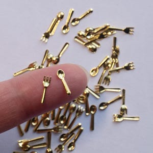 May include: A collection of small, gold-coloured miniature forks and spoons. The tiny utensils are scattered on a white surface, with two pieces held by a finger. The miniature cutlery is likely for dollhouses or craft projects.