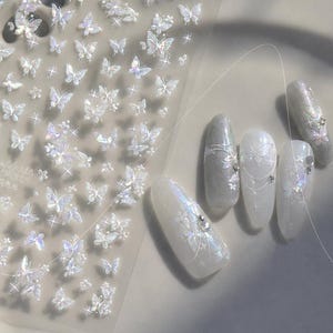 K&ouml;nnte beinhalten: Ein Bogen mit schillernden Schmetterlings-Nagelkunst-Aufklebern neben einem Satz manik&uuml;rter N&auml;gel. Die N&auml;gel sind wei&szlig; und silber lackiert und mit zarten Schmetterlings- und Blumendesigns versehen. Einige N&auml;gel haben kleine, funkelnde Verzierungen.