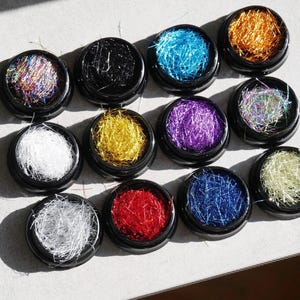 May include: A collection of twelve small, black-lidded jars filled with colorful, shimmering strands. The colors include silver, gold, red, blue, purple, and iridescent hues. These decorative strands are likely used for nail art or craft projects.