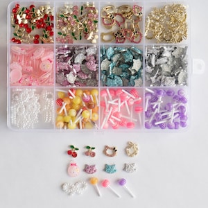 Puede incluir: Un organizador de plástico transparente lleno de varios adornos decorativos. Los compartimentos contienen cerezas rojas, flores doradas, lazos rosas y estrellas plateadas. También se incluyen perlas blancas, piruletas amarillas, rosas y moradas, y varios dijes decorativos.