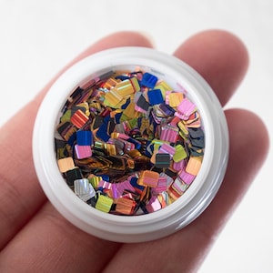 May include: A small, white container filled with colourful, square-shaped glitter. The glitter pieces are a mix of iridescent colours, including gold, blue, pink, and green. The container is held in a hand.