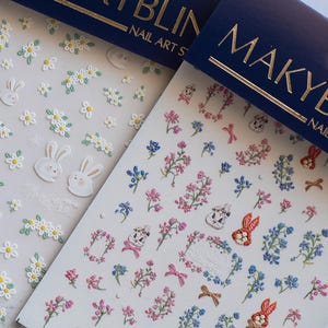 May include: Two sheets of nail art stickers featuring floral and bunny designs. One sheet has white daisies and bunny faces. The other has pink and blue flowers, bows, and bunny illustrations. The brand name "MAKYBLIN" is visible.