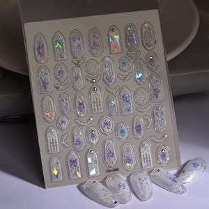 May include: Sheet of iridescent nail art stickers featuring various stained glass window designs with floral accents. The sheet includes multiple shapes and sizes, with the text "Co-249" visible. Several decorated nails are also present.