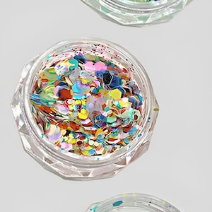 Peut inclure: Trois pots transparents à facettes remplis de paillettes colorées. Les paillettes sont de formes et de tailles variées, dont des hexagones, et dans les tons de bleu, vert, rose, jaune et orange. Les pots sont disposés verticalement sur fond blanc.