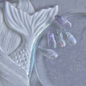 Peut inclure: Un design d'art d'ongle bleu clair sur le th&egrave;me des sir&egrave;nes. Les ongles pr&eacute;sentent des motifs translucides complexes avec des accents argent&eacute;s et des motifs de queue de sir&egrave;ne. Une sculpture de queue de sir&egrave;ne blanche est en arri&egrave;re-plan.