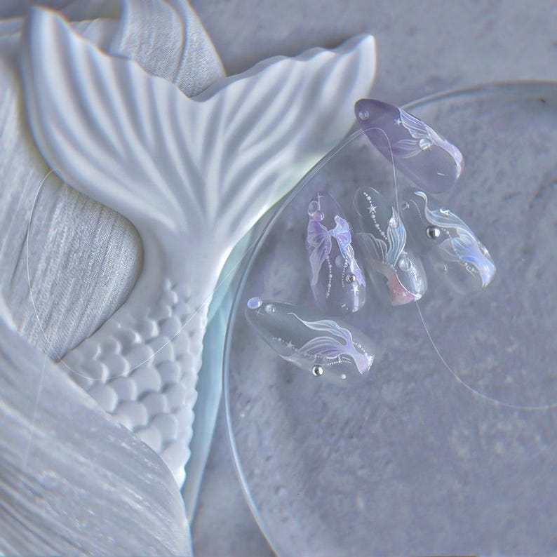 Peut inclure: Un design d'art d'ongle bleu clair sur le th&egrave;me des sir&egrave;nes. Les ongles pr&eacute;sentent des motifs translucides complexes avec des accents argent&eacute;s et des motifs de queue de sir&egrave;ne. Une sculpture de queue de sir&egrave;ne blanche est en arri&egrave;re-plan.