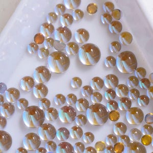 Könnte beinhalten: Eine Sammlung von irisierenden Glascabochons in verschiedenen Größen, von klein bis groß. Die Cabochons sind eine Mischung aus klaren und bernsteinfarbenen Farben mit einem Perlmutt-Finish.
