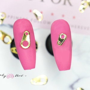 Könnte beinhalten: Zwei rosa Nageldesigns mit goldfarbenen Reizverschlüssen. Die Reize sind wie kleine Reißverschlüsse mit einem kreisförmigen Loch am unteren Ende geformt. Mehrere lose Reize sind um die Nägel herum verstreut.