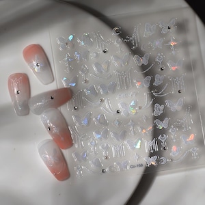 K&ouml;nnte beinhalten: Ein Bogen mit schillernden Nagelkunst-Aufklebern mit Schmetterlings- und Himmelsmotiven. Die Aufkleber sind transparent mit holografischen Akzenten. Mehrere manik&uuml;rte N&auml;gel mit einem Farbverlauf in Pfirsich und Wei&szlig; sind ebenfalls sichtbar, verziert mit kleinen Strasssteinen und Schmetterlingsaufklebern. Auf dem Aufkleberbogen steht der Text "Co-168".