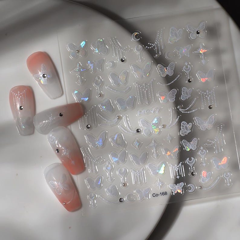 K&ouml;nnte beinhalten: Ein Bogen mit schillernden Nagelkunst-Aufklebern mit Schmetterlings- und Himmelsmotiven. Die Aufkleber sind transparent mit holografischen Akzenten. Mehrere manik&uuml;rte N&auml;gel mit einem Farbverlauf in Pfirsich und Wei&szlig; sind ebenfalls sichtbar, verziert mit kleinen Strasssteinen und Schmetterlingsaufklebern. Auf dem Aufkleberbogen steht der Text "Co-168".