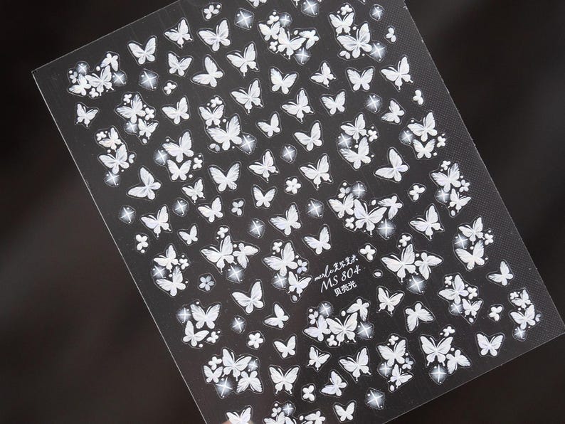 K&ouml;nnte beinhalten: Bogen mit Nagelkunst-Aufklebern mit wei&szlig;en Schmetterlingsdesigns auf schwarzem Hintergrund. Die Aufkleber zeigen Schmetterlinge in verschiedenen Gr&ouml;&szlig;en und einige florale Akzente. Der Text "MS 804" ist sichtbar.